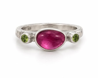 May include: A sterling silver ring with a hammered texture and a 925 hallmark. The ring features a large, oval-shaped, deep pink center stone flanked by two smaller, round, light green peridot stones, all set in smooth silver bezels.