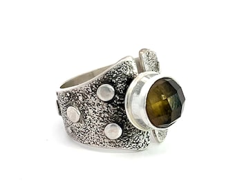 May include: A silver ring with a textured band and four silver dots. The ring features a large, faceted, green and brown gemstone.