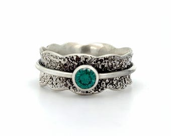 May include: A silver spinner ring featuring a wide band with a dark, heavily textured, oxidized finish and scalloped edges. A smooth, polished silver wire encircles the center, holding a single, round, vibrant green gemstone in a bezel setting.