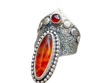 May include: A silver ring with an oval-shaped orange gemstone set in a beaded bezel. The ring has a textured band with small silver dots and a red gemstone at the top.