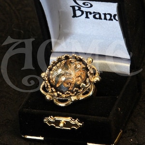 May include: A gold-toned ring with an ornate design, featuring a central dark stone with intricate patterns. The ring is displayed in an open black velvet box with gold accents. The box has the word "Brand" printed on the inside.