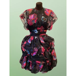 Könnte beinhalten: Schwarzes Kleid mit floralem Muster in Rosa-, Blau- und Lilatönen. Das Kleid hat einen transparenten Überzug auf dem Oberteil und den Ärmeln, eine taillierte Taille und einen geschichteten Rock. Das Kleid wird auf einer Schaufensterpuppe präsentiert.