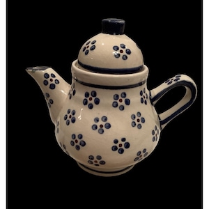 May include: A ceramic teapot with a lid, featuring a white base adorned with blue floral patterns and small red accents. The handle and spout are integrated into the design, with a dark blue trim. The teapot is designed for brewing tea.