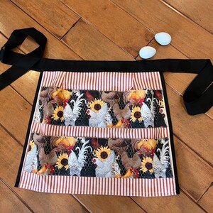 May include: A black and white apron with a red and white striped border. The apron has three pockets and features a pattern of chickens and sunflowers.
