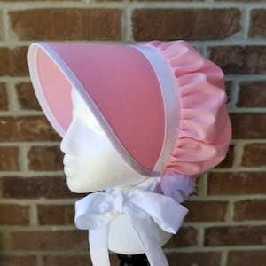 May include: A pink bonnet with a wide brim and a white ribbon tie. The bonnet features a rounded crown and a white trim along the edge of the brim. The bonnet is displayed on a white mannequin head against a brick background.