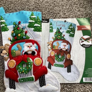 Könnte beinhalten: Zwei Weihnachtsstrümpfe aus Filz. Einer ist fertig und zeigt den Weihnachtsmann in einem roten Lastwagen mit einem Kranz und dem Text "B-JOLLY". Der andere ist ein Bausatz mit dem Namen "Lucas" und den Worten "Jolly Deliveries".