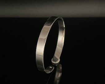 Op de afbeelding: Een gepolijste zilveren manchetarmband. De open armband heeft een plat, rechthoekig profiel en een glad, reflecterend oppervlak. Het sieraad staat tegen een zwarte achtergrond, wat de metaalglans benadrukt. Een eenvoudig, elegant accessoire.