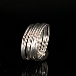 May include: A silver-colored ring with multiple bands wrapped around each other. The ring is made of thin, shiny metal and has a simple, elegant design. The ring is shown against a black background.