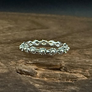 Peut inclure: Une bague en argent avec un motif floral. La bague présente un motif répété de petites fleurs détaillées autour de l'anneau. La bague est présentée sur une surface en bois.