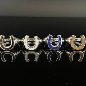 May include: Five silver horseshoe rings with different colored inlays. The rings are arranged in a row from left to right. The inlays are black, white, blue, brown, and gold.