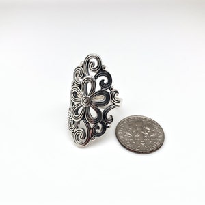 May include: A silver-colored ring with a floral design. The ring features a central flower shape surrounded by swirling, openwork patterns. A U.S. dime is next to the ring for size comparison. The ring is on a white background.