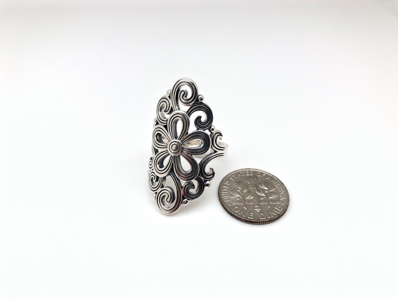 May include: A silver-colored ring with a floral design. The ring features a central flower shape surrounded by swirling, openwork patterns. A U.S. dime is next to the ring for size comparison. The ring is on a white background.