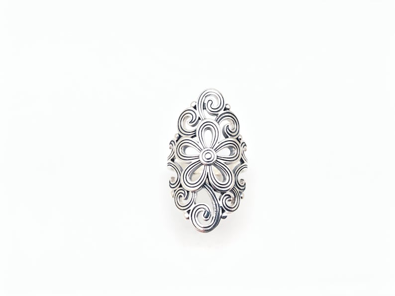 May include: A silver-colored ring with a floral design. The ring features a central flower with a circular center and petals, surrounded by swirling, openwork patterns. The ring is set against a white background.