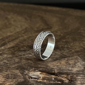 Puede incluir: Un anillo de plata con un diseño trenzado. El anillo está sobre una superficie de madera.