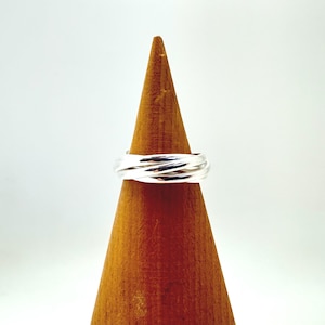 Könnte beinhalten: Ein Ring aus poliertem Silber, der aus drei ineinandergreifenden Bändern besteht, die sich umeinander winden und ein einziges, durchgehendes Stück bilden. Der Ring wird auf einem glatten, hellbraunen Holzkegel vor weißem Hintergrund präsentiert.