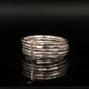 May include: A silver ring with a textured, hammered design. The ring features multiple bands stacked together, with a vertical bar connecting them. The ring is displayed on a reflective black surface, creating a mirrored effect.