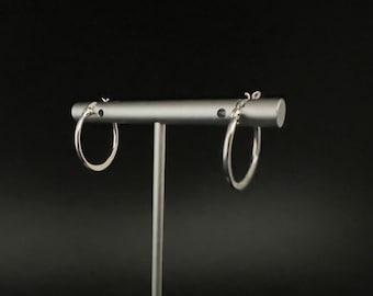 May include: A pair of silver hoop earrings displayed on a stand. The earrings are simple and circular in shape.