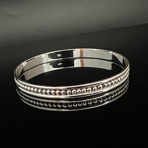 May include: A shiny silver bangle bracelet with a polished finish. The bracelet features a row of small, evenly spaced beads between two smooth bands. The jewelry is displayed on a reflective black surface, highlighting its bright, metallic appearance.