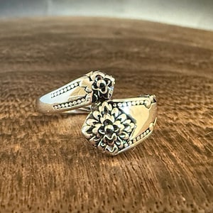 May include: Two silver rings with floral designs. One ring is in focus, displaying a detailed flower pattern on the face and dotted accents along the band. The other ring is slightly out of focus, showing a similar design. The rings are set against a wooden background.