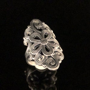 May include: A silver-colored ring with a floral design. The ring features a large flower in the center, surrounded by swirling, openwork patterns. The ring is set against a black background.