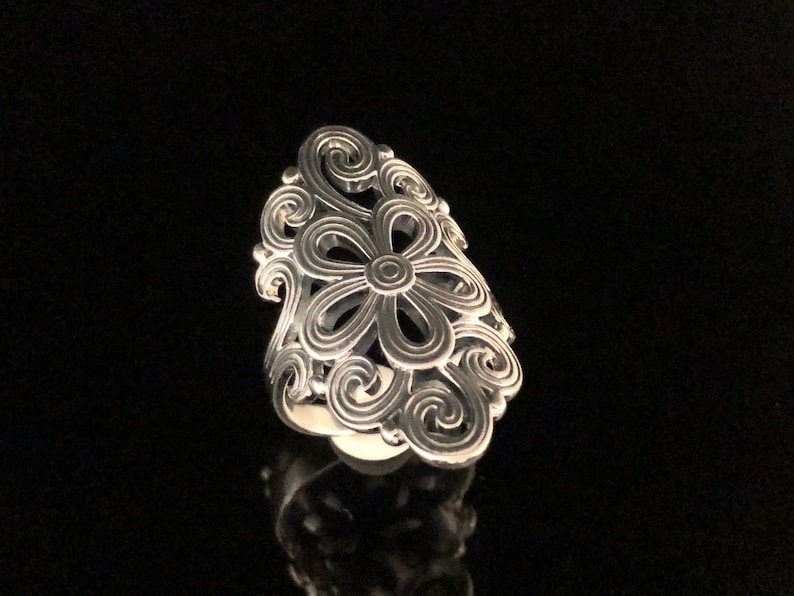 May include: A silver-colored ring with a floral design. The ring features a large flower in the center, surrounded by swirling, openwork patterns. The ring is set against a black background.