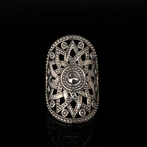 May include: Silver-toned statement ring with an intricate openwork design. The ring features a central circular element surrounded by a starburst pattern, with detailed filigree and dot accents. The elongated oval shape adds a bold, eye-catching element.