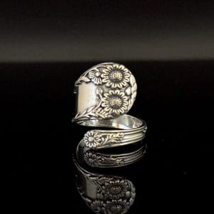Puede incluir: Un anillo de cuchara de plata con un diseño floral. El anillo presenta un patrón detallado de flores y hojas, con un diseño floral prominente en la cara del anillo. El anillo se muestra sobre un fondo negro.