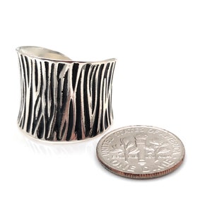 Könnte beinhalten: Ein breiter Silberring mit einem schwarz-weißen Zebrastreifenmuster. Der Ring wird neben einer 10-Cent-Münze zur Größenvergleich gezeigt.