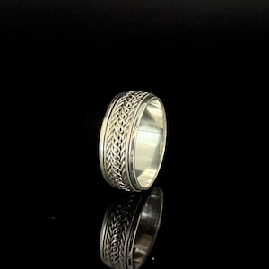May include: A silver ring with a woven design around the band. The ring is highly reflective, with a polished finish. The ring is set against a black background, with its reflection visible below. Jewelry.