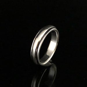 Könnte beinhalten: Ein silberner Ring mit einer glatten, abgerundeten Oberfläche und einem leicht erhöhten Rand. Der Ring ist schlicht und elegant.