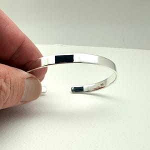 May include: A polished silver cuff bracelet with a rectangular black inlay. The open-ended design allows for easy wear. The bracelet is a simple, elegant piece of jewelry.