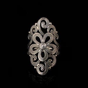 May include: A silver-colored ring with an openwork floral design. The ring features a central flower shape surrounded by swirling, ornate patterns. The ring is set against a black background, highlighting its intricate details.
