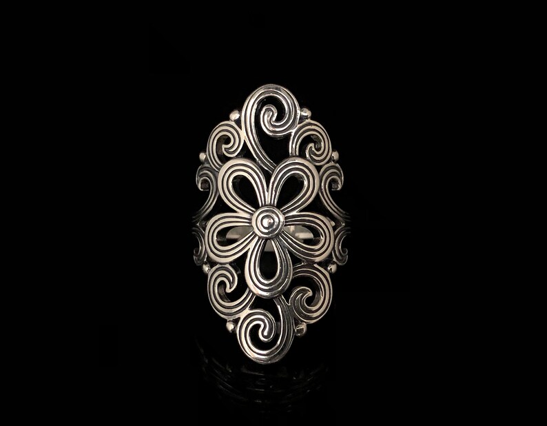 May include: A silver-colored ring with an openwork floral design. The ring features a central flower shape surrounded by swirling, ornate patterns. The ring is set against a black background, highlighting its intricate details.