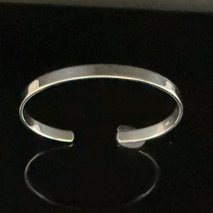 May include: A silver cuff bracelet with a simple, open design. The polished metal reflects light, creating a bright appearance. The bracelet is shown against a dark background, highlighting its shape and shine.