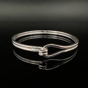 May include: A shiny silver bangle bracelet with a hook closure. The bracelet is round and has a smooth, polished surface. The hook closure is a unique design element. The bracelet is displayed on a reflective black surface.