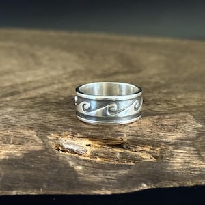May include: A silver ring with a wave pattern. The ring has a band decorated with a series of stylised waves, evoking a nautical or ocean theme. The ring is made of a shiny, metallic material, and the waves are etched into the band.