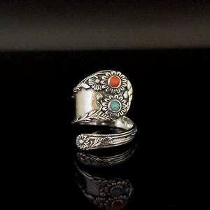 Puede incluir: Un anillo de plata con forma de cuchara con detalles florales y de hojas. El anillo presenta dos incrustaciones circulares, una roja y otra turquesa. El anillo se muestra sobre una superficie negra.