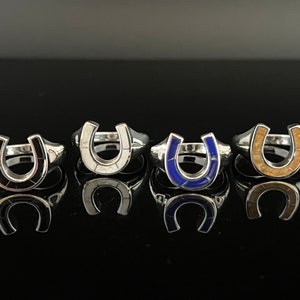 May include: Four silver horseshoe rings with different colored inlays. The rings are arranged in a row on a black surface. The inlays are black, white, blue, and brown.