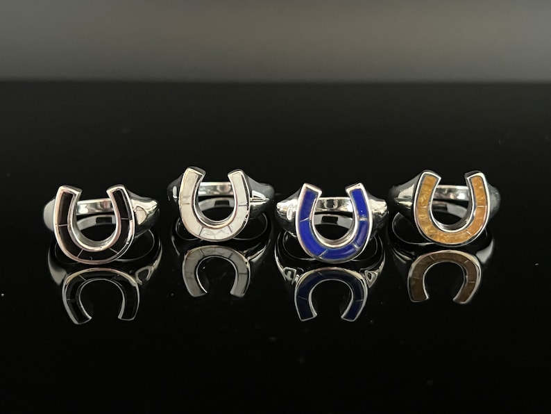 May include: Four silver horseshoe rings with different colored inlays. The rings are arranged in a row on a black surface. The inlays are black, white, blue, and brown.