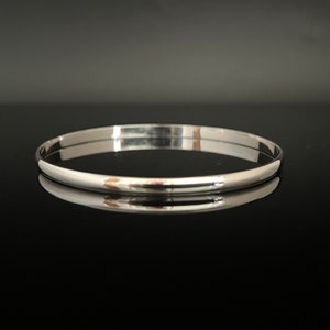 May include: A polished silver bangle bracelet. The smooth, rounded design reflects light, creating a bright, shiny appearance. The bracelet is a simple, elegant piece of jewellery. The bangle is approximately 0.6 cm wide.