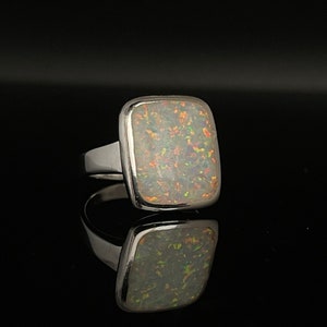 May include: A silver ring featuring a large, square opal gemstone. The opal exhibits a vibrant play of colour with flashes of orange, green, and yellow against a white base. The ring's band is thick and polished, reflecting the dark background.