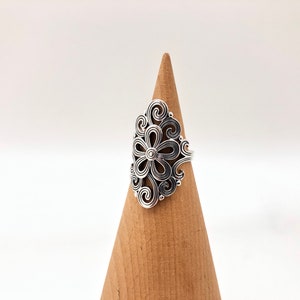 May include: A silver-colored ring with a floral design. The ring features a central flower shape surrounded by intricate swirling patterns. The ring is displayed on a wooden cone against a white background.