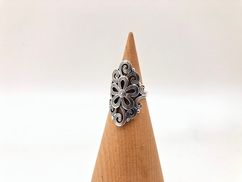 May include: A silver-colored ring with a floral design. The ring features a central flower shape surrounded by intricate swirling patterns. The ring is displayed on a wooden cone against a white background.