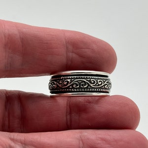 May include: A silver ring with an ornate, swirling pattern around the band. The ring is held between fingers against a white background. The design features a series of interconnected curves and dots, creating a detailed, textured appearance.