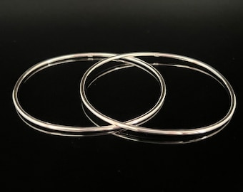 Può includere: Due braccialetti in argento, uno leggermente più grande dell'altro, sono intrecciati e giacciono su una superficie nera.