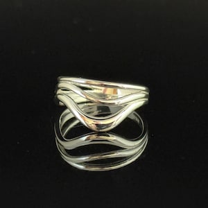 May include: A silver ring with a unique, layered design. The ring features multiple wavy bands that create an open, flowing aesthetic. The polished metal reflects light, highlighting the ring's curves and contours. The ring is a piece of jewelry.