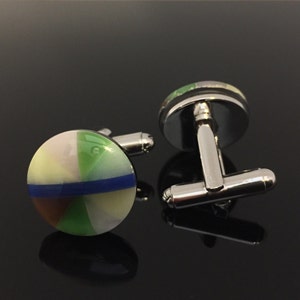May include: A pair of round cufflinks with a colorful design. Each cufflink features a silver-tone metal base and a circular face divided into segments of white, green, brown, and blue. The cufflinks are displayed on a reflective black surface.
