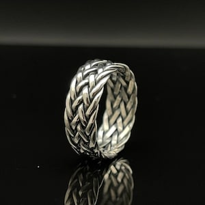 May include: A silver-colored ring with a woven design. The ring's band features a detailed pattern of interwoven strands, creating a textured surface. The ring is displayed against a dark background, highlighting its metallic sheen.