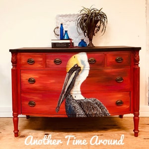 May include: A red painted dresser with a pelican painted on the front. The dresser has six drawers with brass pulls. The dresser is on a wooden floor. The text "Another Time Around" is written below the dresser.