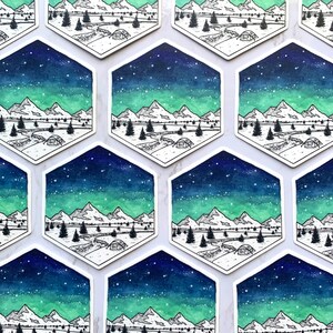 May include: A set of nine hexagonal stickers featuring a watercolor illustration of a mountain range under a starry sky with a green aurora borealis. The illustration is in black and white with a blue and green watercolor wash.